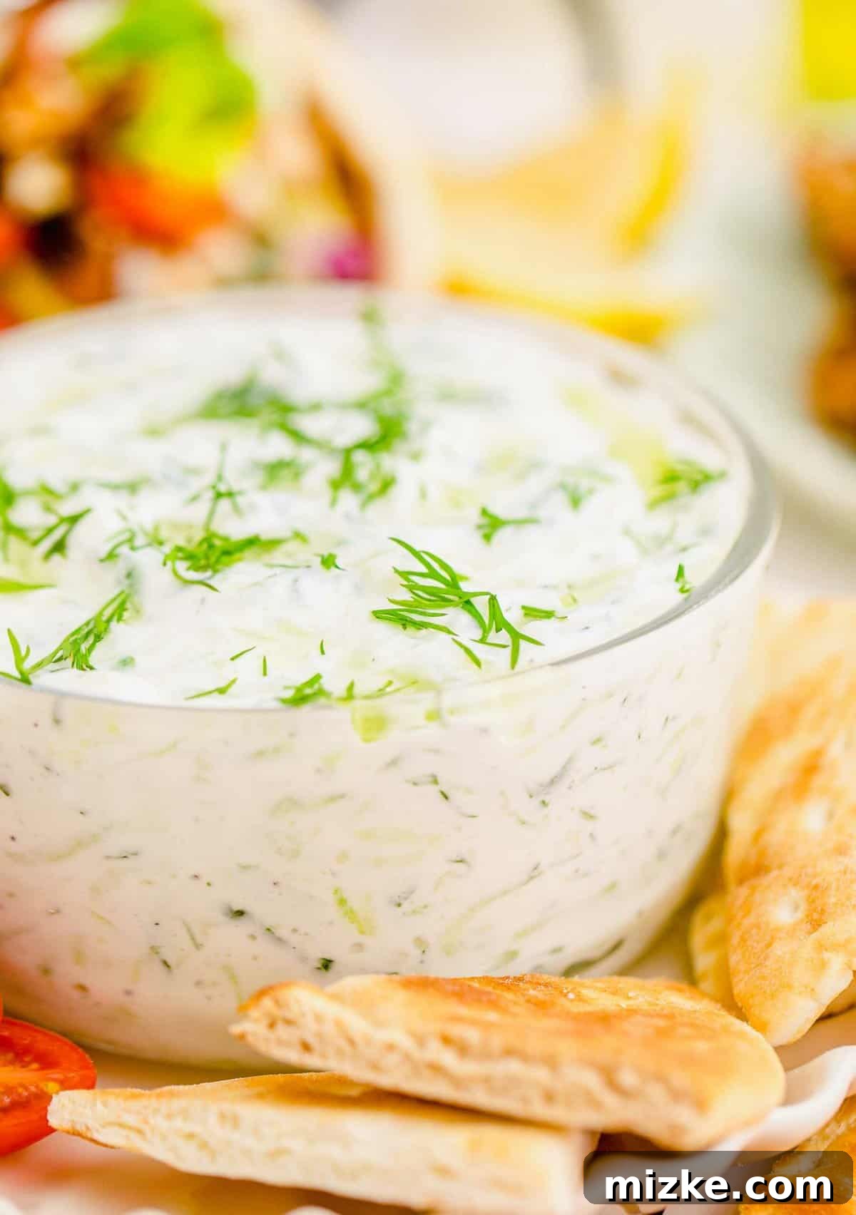 Creamy Homemade Tzatziki 4 A close-up view of a glass bowl generously filled with creamy homemade tzatziki dip, elegantly garnished with fresh dill and a swirl of golden olive oil, surrounded by crisp pita chips ready for dipping.