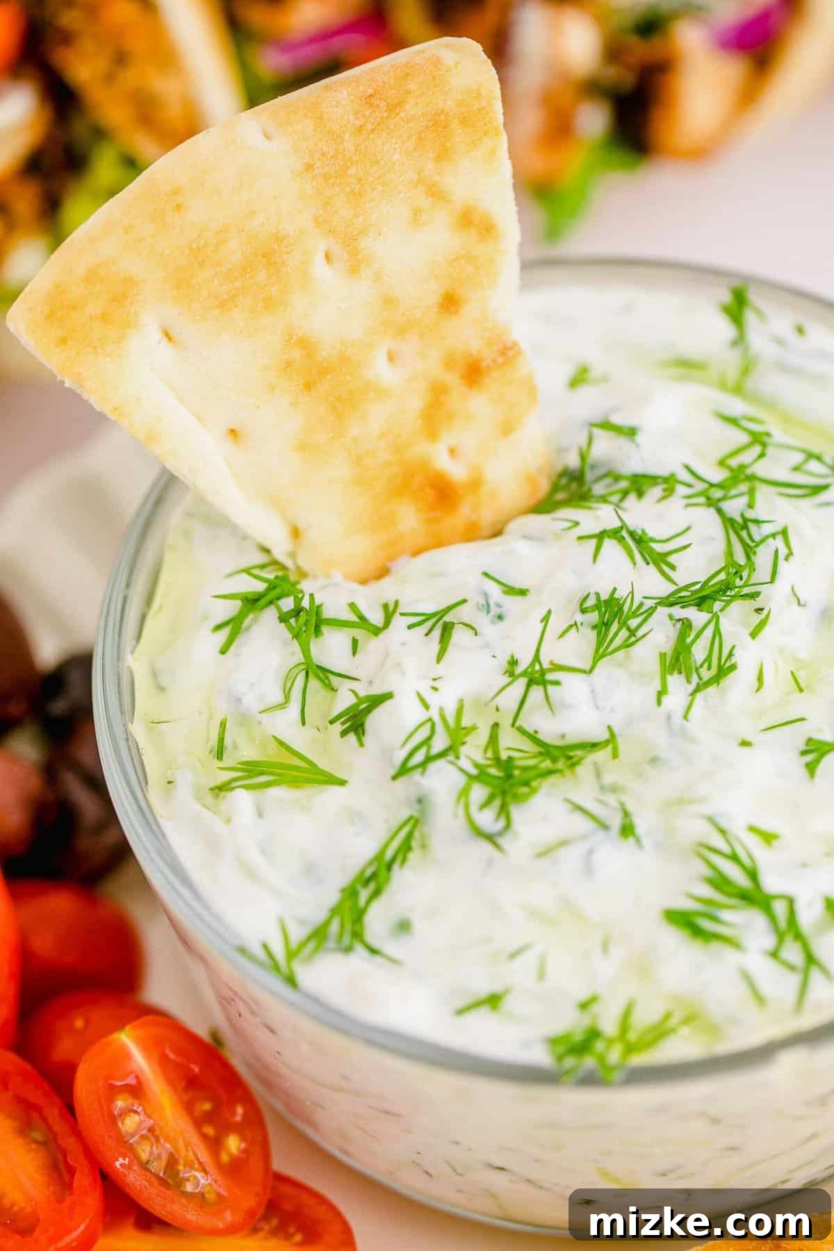 Creamy Homemade Tzatziki 5 A golden pita chip emerging from a luscious bowl of creamy homemade tzatziki dip, exquisitely adorned with fresh dill and a light drizzle of olive oil, irresistibly inviting a taste.