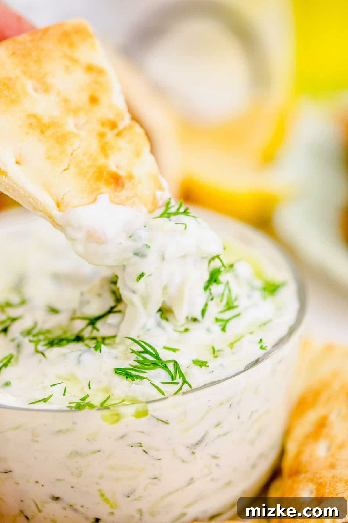Creamy Homemade Tzatziki 6 A hand gracefully dipping a golden pita chip into a luscious bowl of homemade tzatziki, perfectly highlighting the dip's creamy texture and inviting appeal.