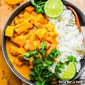 A black bowl filled with white rice and a generous serving of vibrant chicken pumpkin curry, garnished with fresh cilantro and lime wedges.