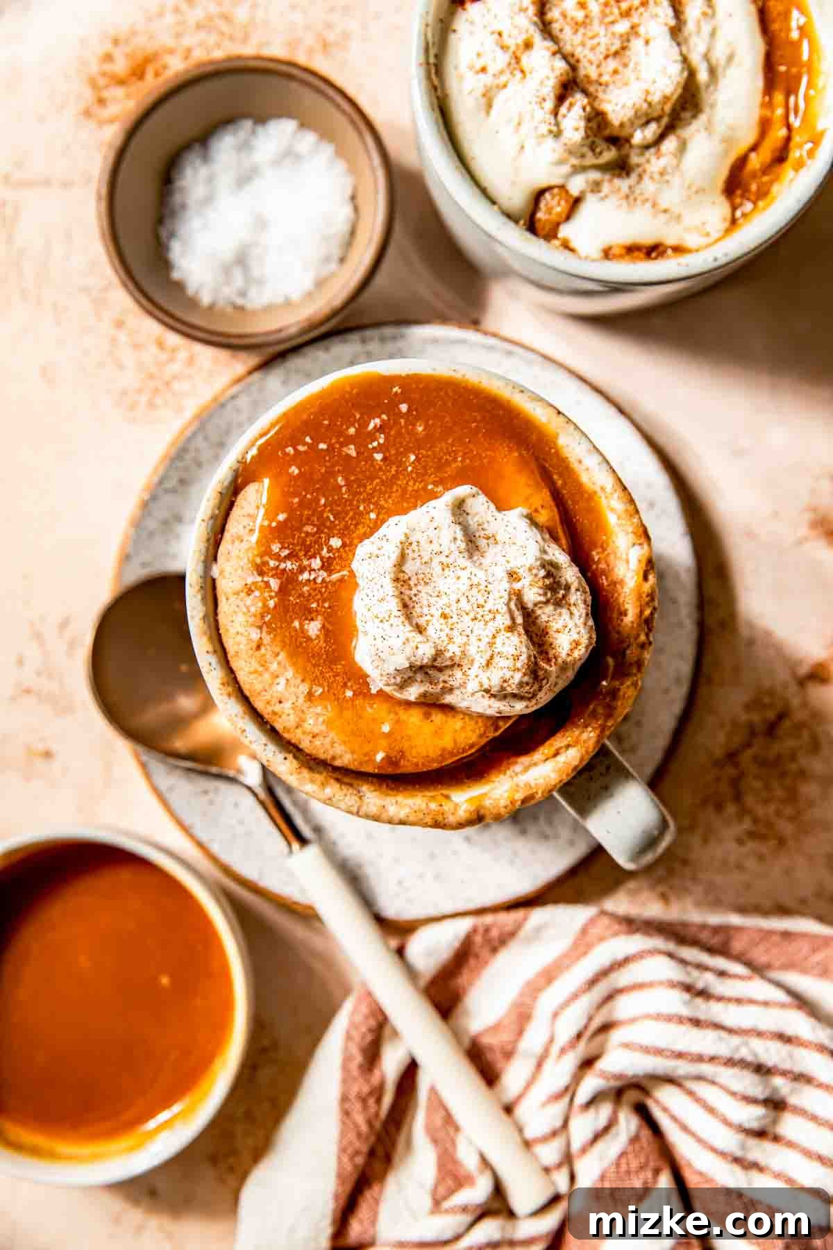 A mug of salted caramel apple mug cake topped with whipped cream and cinnamon sits on a plate with a spoon; a bowl of salted caramel sauce, a bowl of salt, and a striped napkin are nearby, evoking a warm autumn scene.