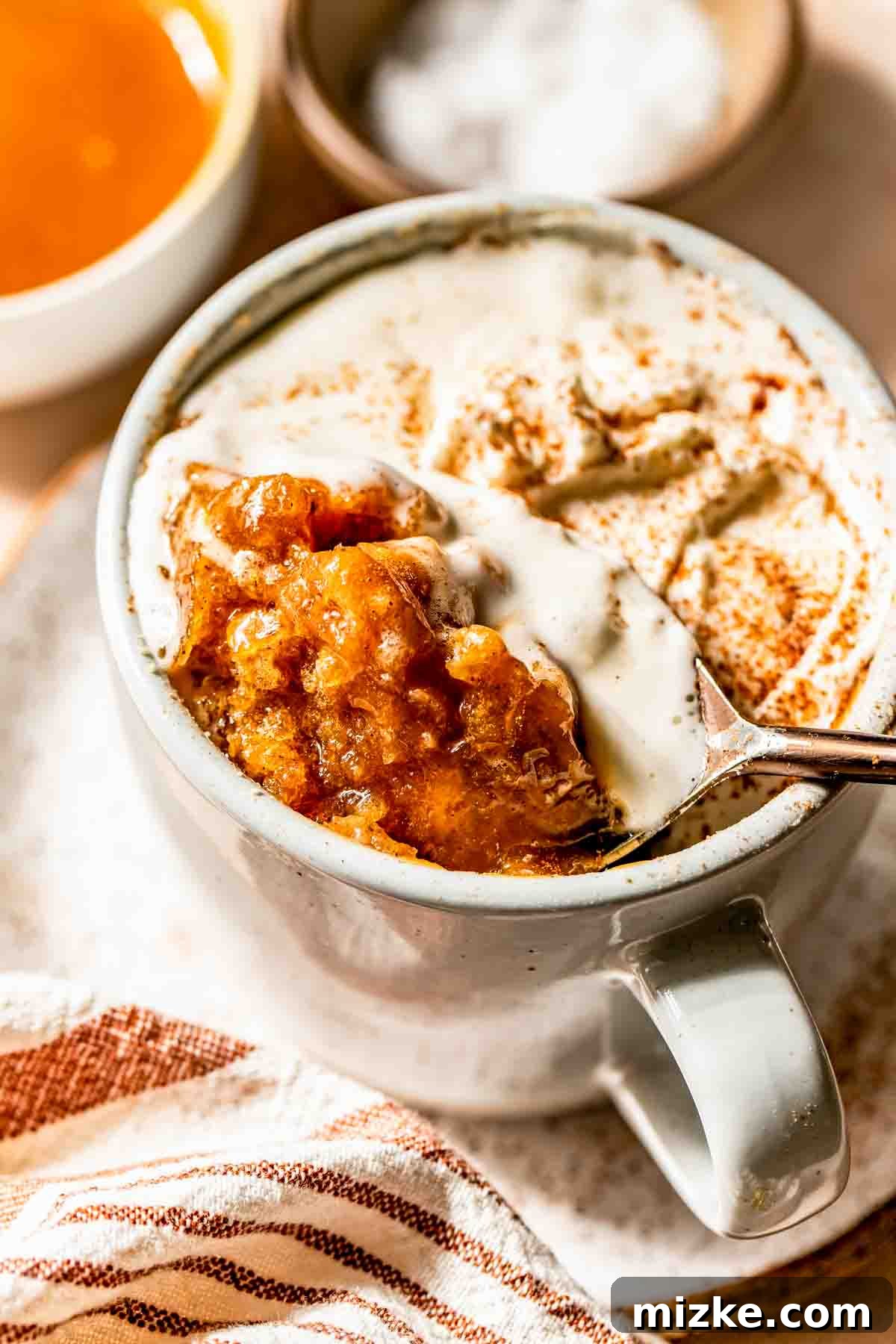 A mug filled with a baked salted caramel apple dessert topped with creamy sauce, with a spoon lifting a portion; a striped napkin and small bowls are nearby, suggesting an inviting dessert setup.
