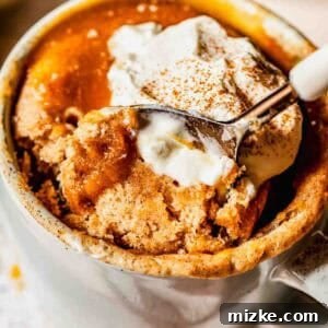 A close-up of a mug filled with cinnamon spiced mug cake, topped with a dollop of whipped cream and a sprinkle of cinnamon, with a spoon scooping out a portion.