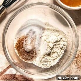 A glass bowl with flour, sugar, cinnamon, and salt on a countertop, next to a whisk, a bowl of salted caramel sauce, and a striped towel, showcasing the dry ingredients.