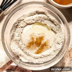 A glass bowl with flour, sugar, oil, vanilla extract, and milk, next to a bowl of salted caramel sauce and a whisk on a light countertop with a striped towel, illustrating the mixing of wet and dry ingredients.
