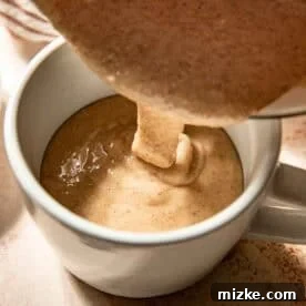 Thick, cinnamon-spiced batter being poured from a bowl into a white mug on a light surface, showing the transfer to the mug.