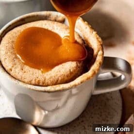 A spoon pours caramel sauce over a freshly cooked mug cake in a white mug placed on a speckled plate with a spoon beside it, ready to be served.