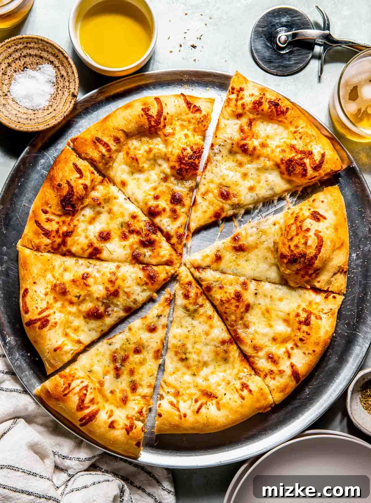 Bianco Bliss 2 Perfectly baked white pizza sliced into eight pieces on a metal tray, with a pizza cutter, bowls of salt and olive oil, and a striped kitchen towel nearby.