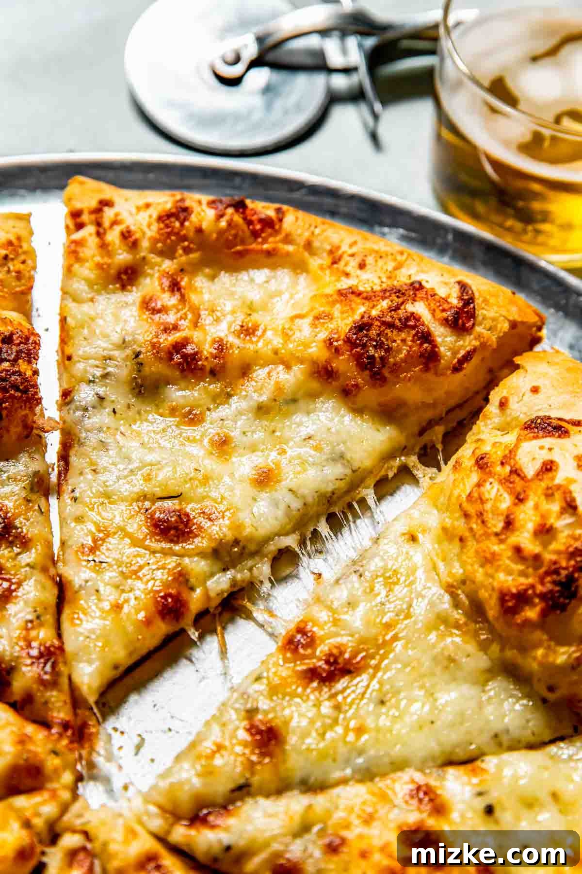 Bianco Bliss 4 Close-up view of a freshly baked white pizza, perfectly sliced on a metal tray, with a pizza cutter and a glass of beer subtly in the background.