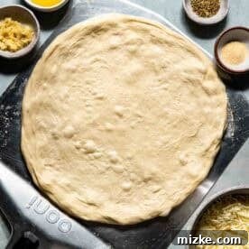 Raw pizza dough on a peel surrounded by small bowls containing garlic, spices, shredded cheese, salt, and olive oil on a light surface, ready for toppings.