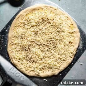 Unbaked pizza dough topped with shredded mozzarella cheese on a pizza peel, with a brush and small bowls of ingredients on a gray surface.