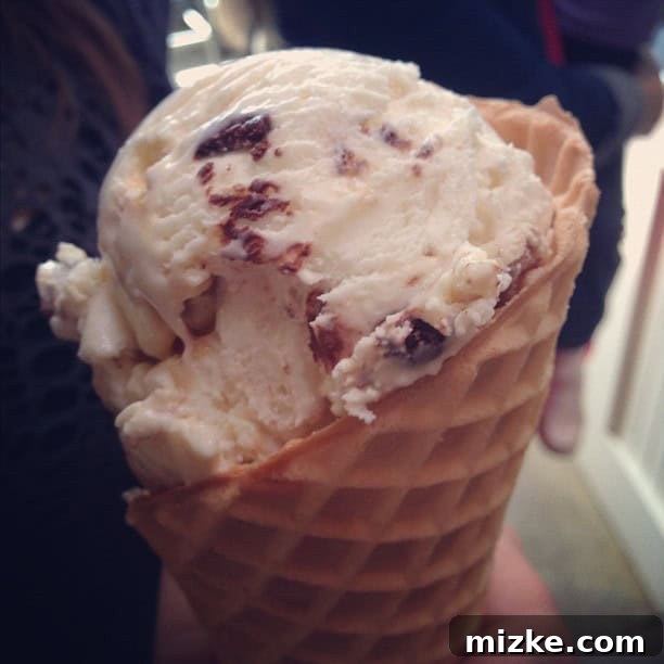 A hand holding a scoop of Salt & Straw ice cream, showing off a unique flavor with visible ingredients, against a blurred background.