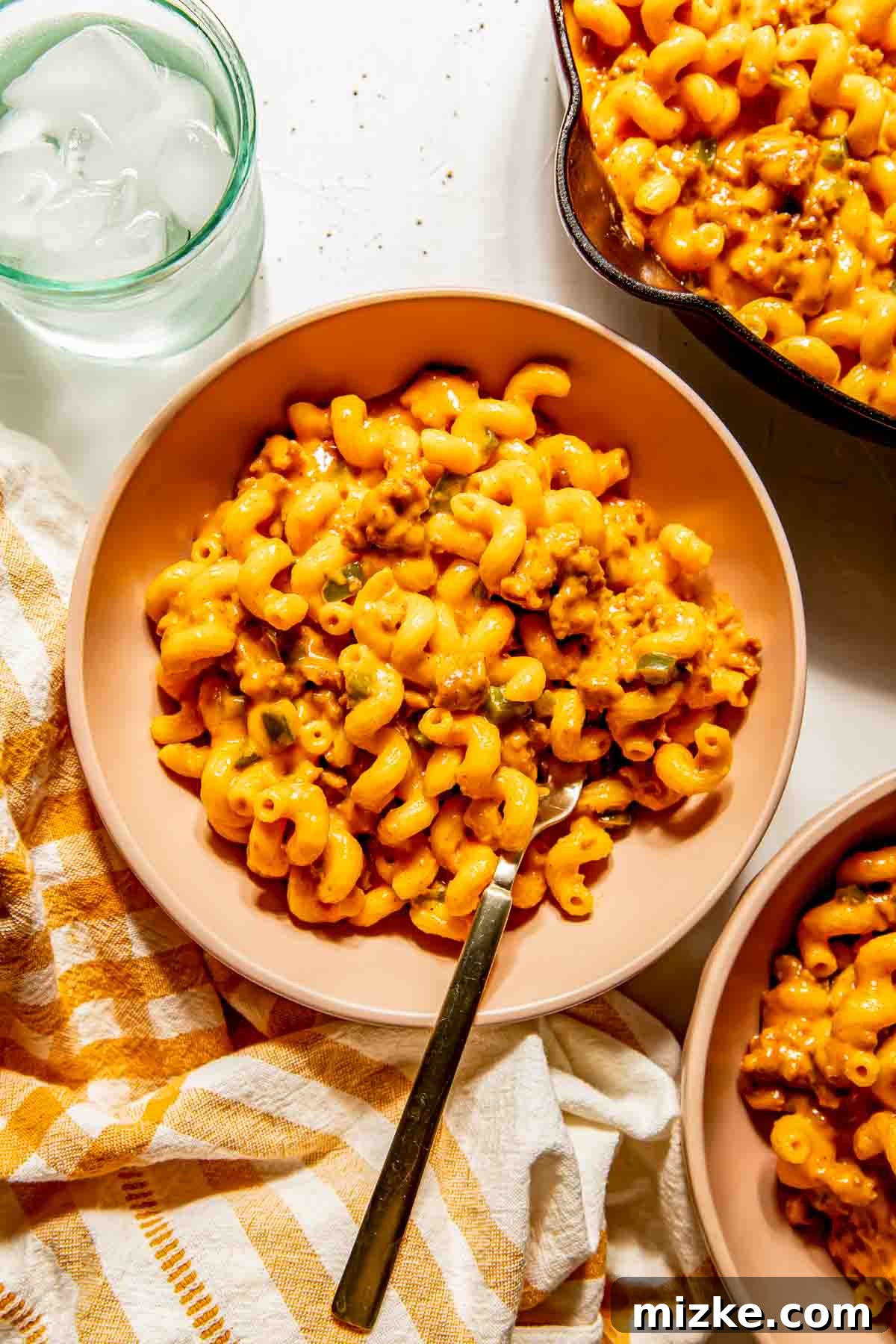 Smoky Chorizo Mac and Cheese 4 A ceramic bowl of steaming chorizo macaroni and cheese, adorned with fresh herbs, sits on a striped kitchen towel, with a fork ready to dig in and a glass of ice water nearby.
