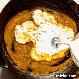 Smoky Chorizo Mac and Cheese 8 Milk is being poured into a cast iron skillet containing a butter and flour mixture. A checkered cloth is partially visible at the bottom of the image.