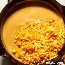 Smoky Chorizo Mac and Cheese 9 A cast iron skillet filled with creamy cheese sauce and topped with a pile of shredded cheddar cheese, next to a checkered kitchen towel.