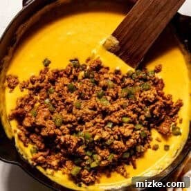 Smoky Chorizo Mac and Cheese 10 A cast iron skillet filled with melted cheese sauce being mixed with cooked ground meat and diced green peppers using a wooden spatula.