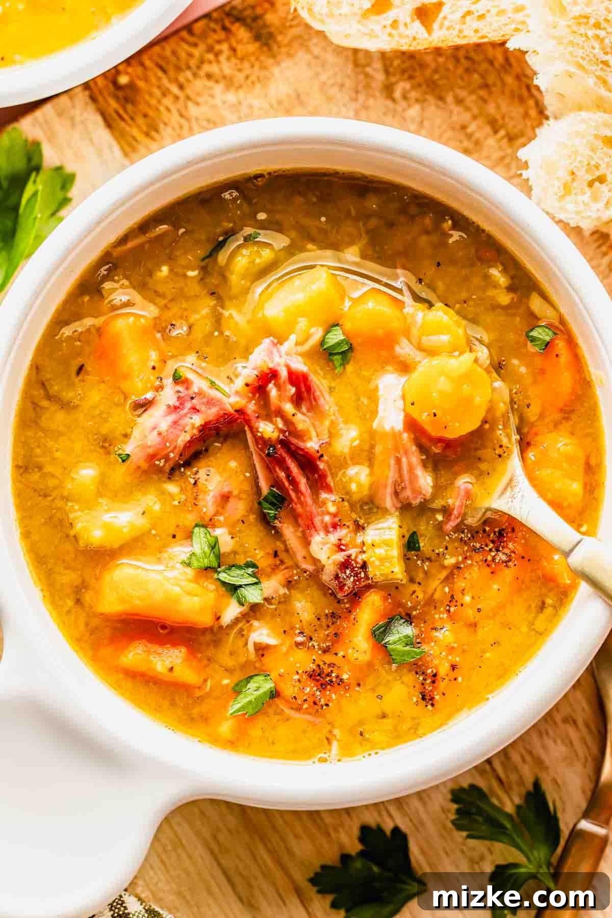 Hearty Slow Cooker Ham and Split Pea Soup 3 A close-up shot of a bowl of split pea soup with shredded ham, vibrant carrots, and fresh herbs, served with a spoon on a rustic wooden surface next to crusty bread slices and parsley sprigs.