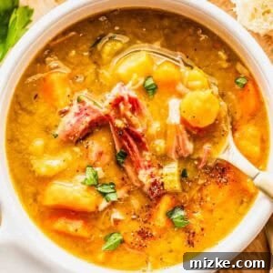 A bowl of split pea soup with shredded ham, carrots, and herbs, served with a spoon on a wooden surface next to bread slices and parsley.