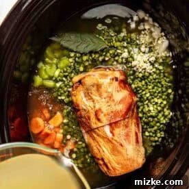 A slow cooker filled with a ham shank, split green peas, chopped carrots, celery, minced garlic, a bay leaf, and broth being poured in.