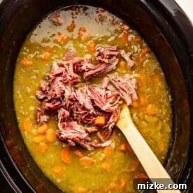 A slow cooker filled with split pea soup, visible carrots, and shredded ham being stirred with a wooden spoon.