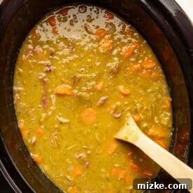 A slow cooker filled with split pea soup containing chopped carrots and ham, with a wooden spoon resting inside.