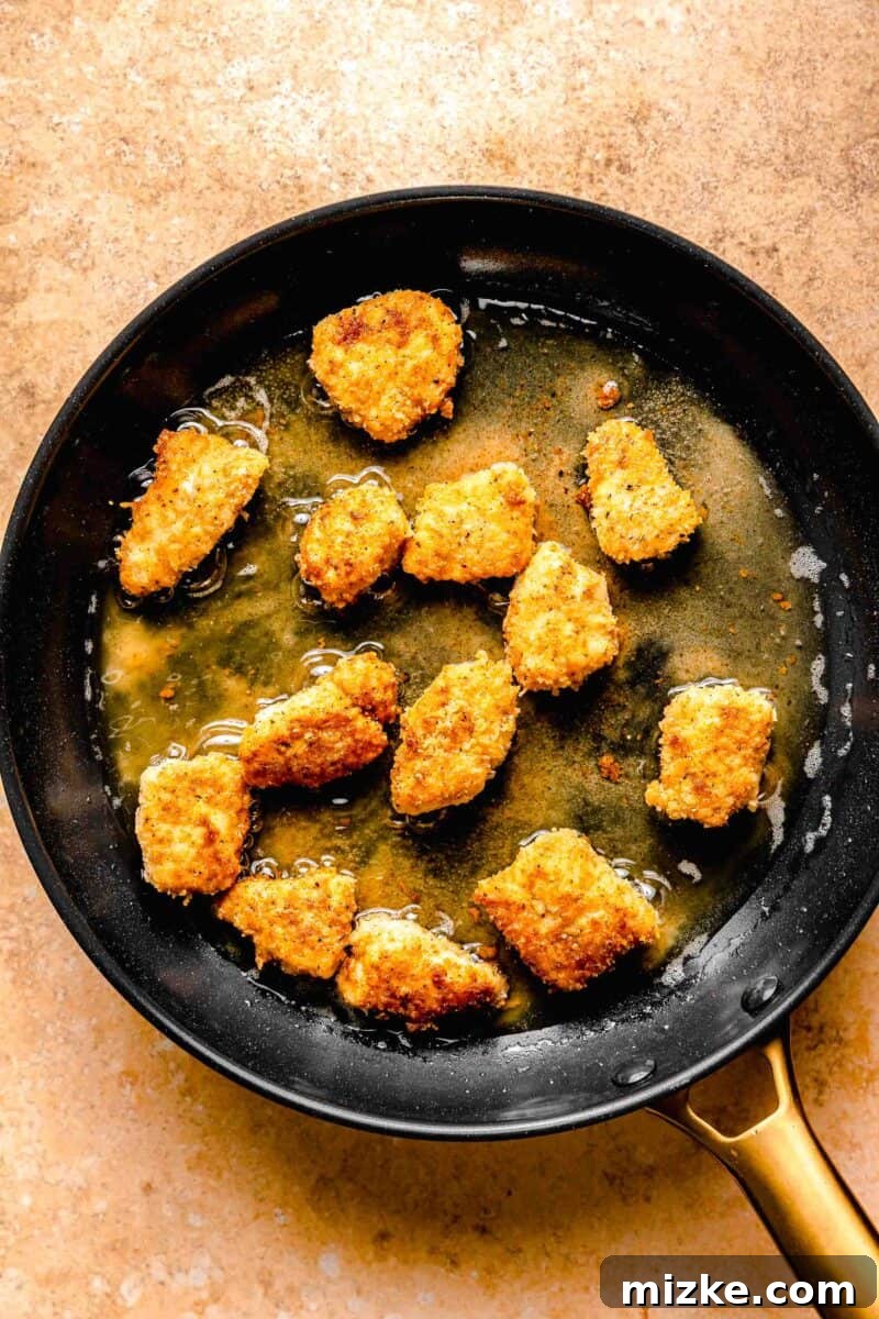 Perfectly crispy cubes of breaded chicken are coated in a delicious spicy sauce that fills every nook and cranny in this bang bang chicken. Golden-brown, crispy breaded chicken cubes frying in hot oil in a skillet, showcasing the cooking process before sauce application.