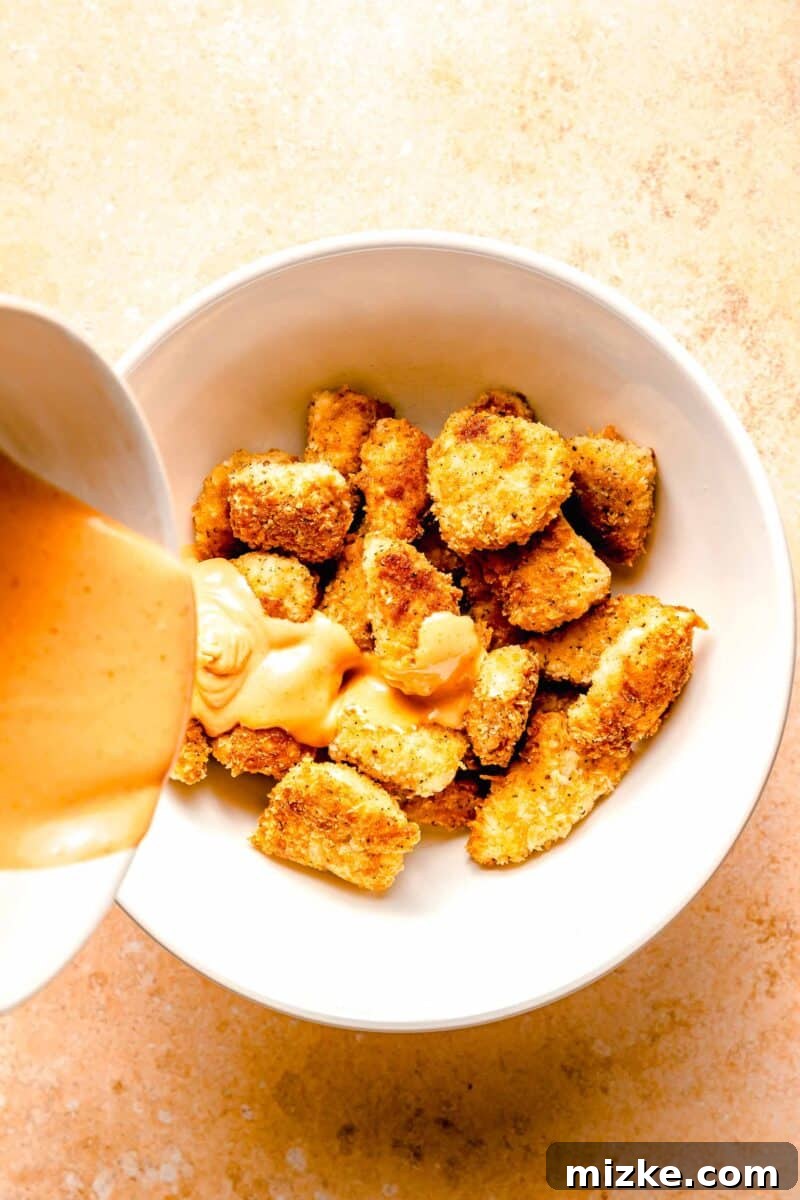 Perfectly crispy cubes of breaded chicken are coated in a delicious spicy sauce that fills every nook and cranny in this bang bang chicken. A large serving bowl filled with golden-brown fried chicken pieces, with a rich, creamy Bang Bang sauce being poured generously over them, ready for tossing and serving.
