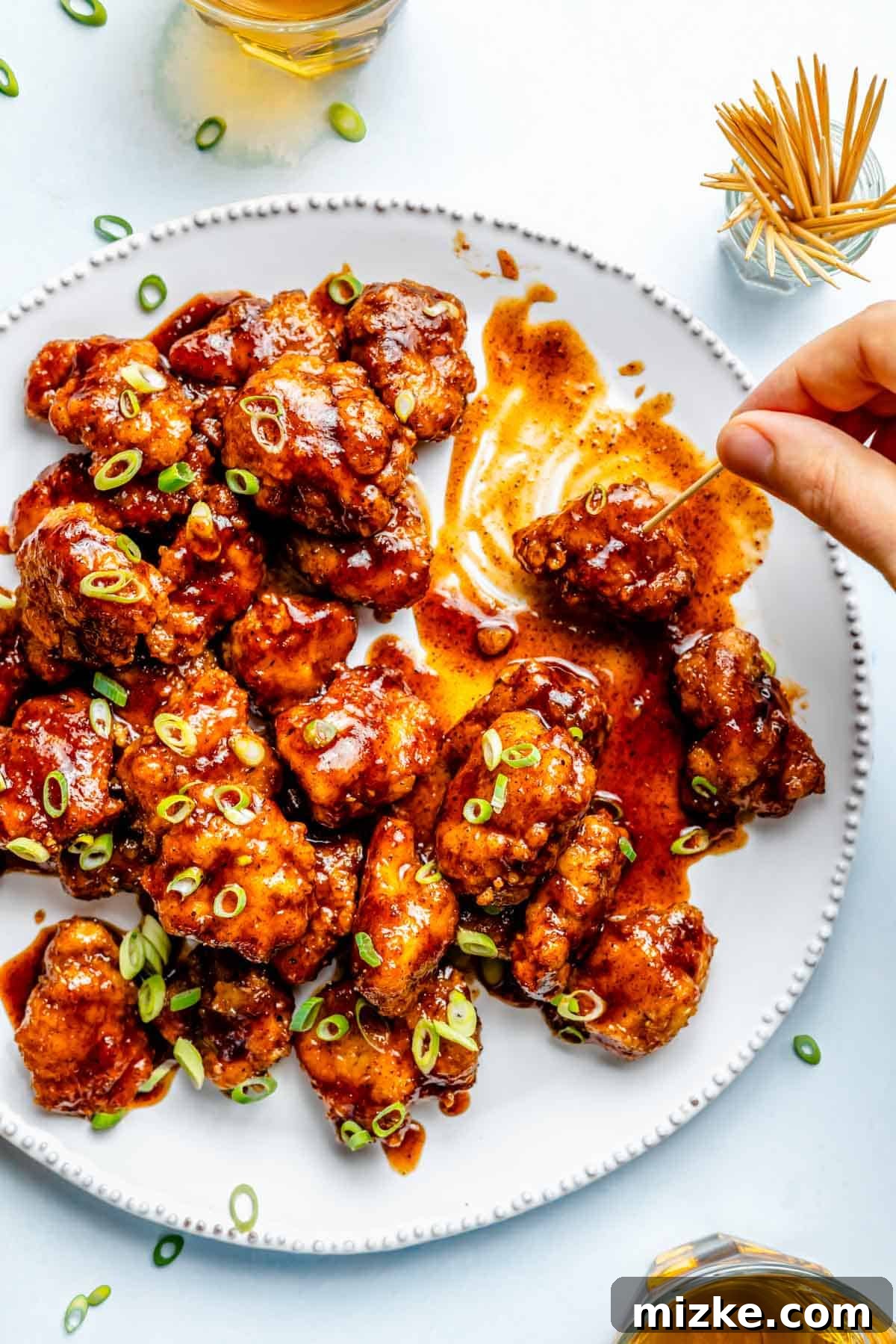 Chipotle Honey Glazed Chicken Bites 3 A close-up view of golden brown Chipotle Honey Chicken Poppers on a white platter, with a hand delicately picking one up using a toothpick, emphasizing their perfect size.