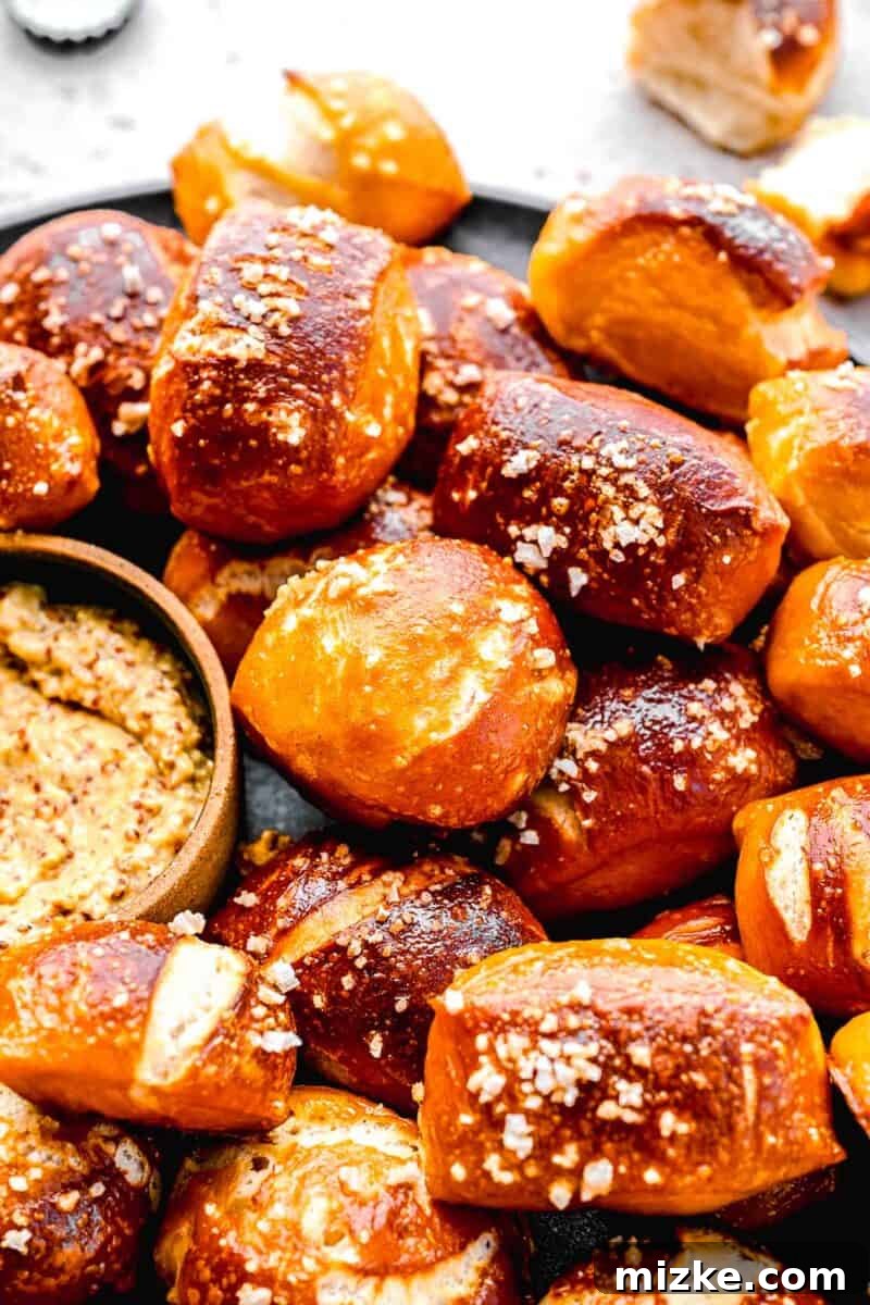 A close-up view of golden-brown pretzel bites arranged on a plate, featuring the textured coarse sea salt, next to a small bowl of whole-grain mustard.