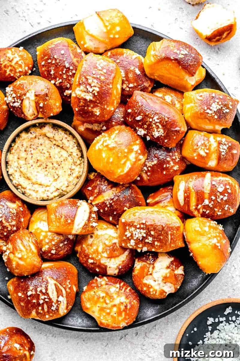 A white serving plate filled with numerous golden-brown pretzel bites, artfully arranged next to a small bowl of whole grain mustard on a light background.
