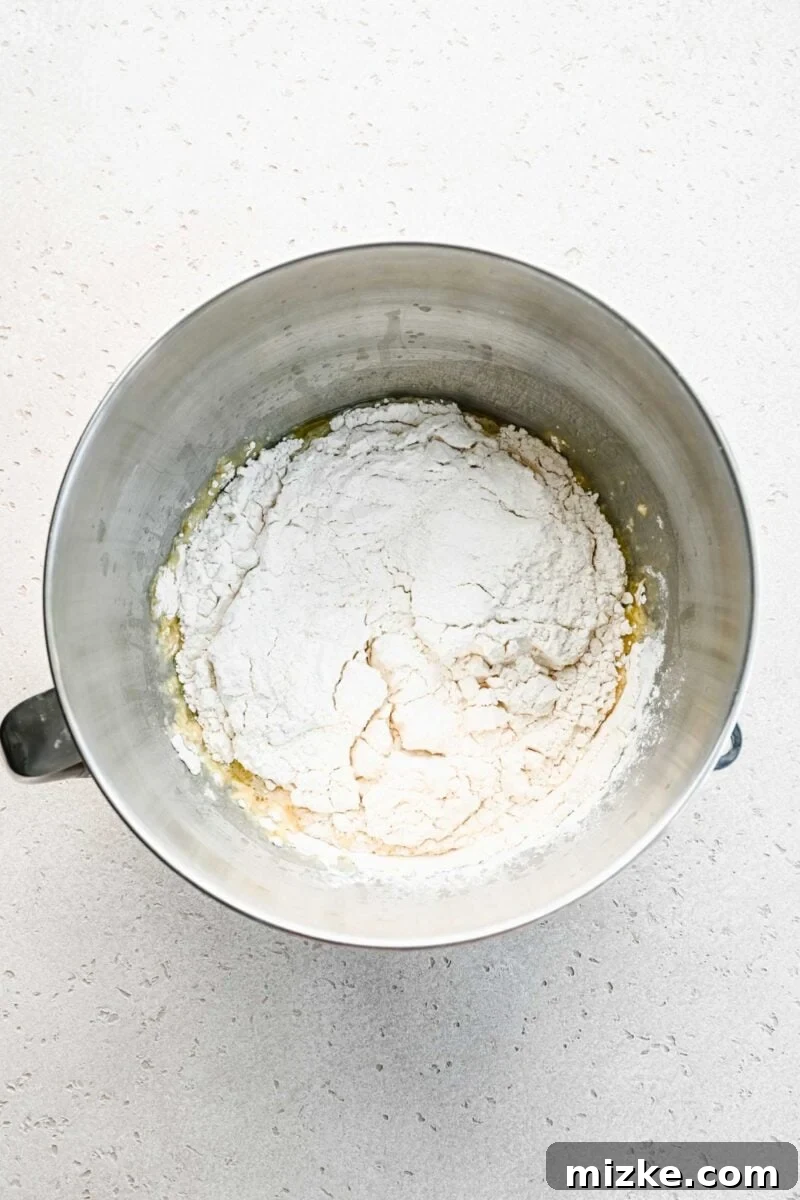 Dry ingredients, specifically flour, being added to the wet mixture in a stand mixer to form the pretzel dough.
