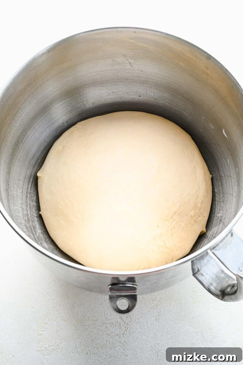 Pretzel dough, visibly risen and doubled in size, in a mixing bowl covered with plastic wrap.