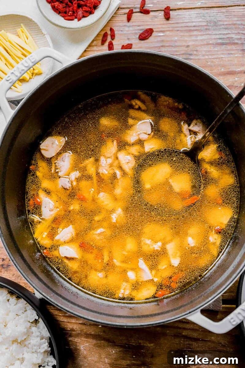 Soul-Warming Rice Wine Chicken Soup 3 Chinese chicken noodle soup in a large pot with a ladle. Small bowls of ginger, goji berries, and white rice surround the pot, highlighting fresh ingredients.