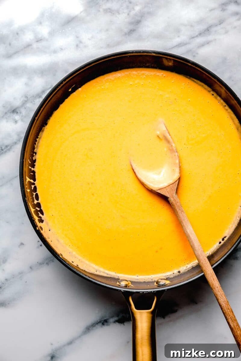 Kickin' Buffalo Mac and Cheese 5 A creamy, golden cheese sauce is being stirred in a skillet with a wooden spoon, showcasing its smooth, rich consistency.