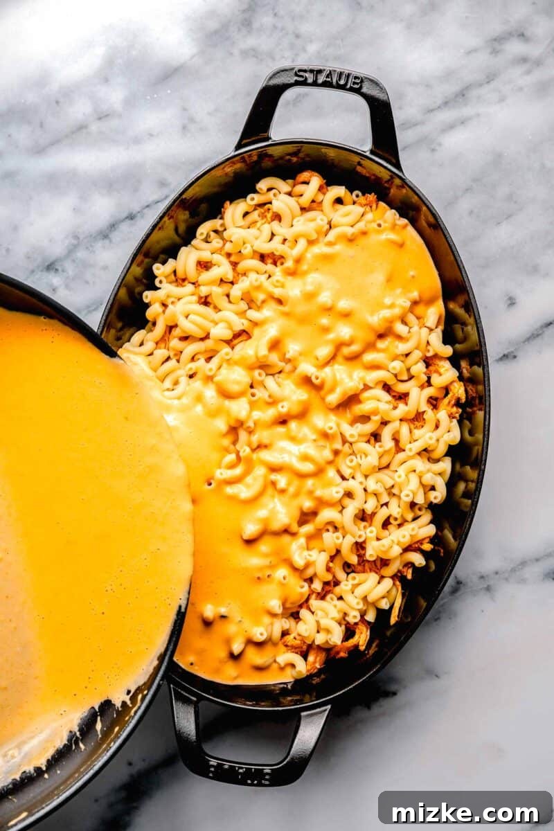 Kickin' Buffalo Mac and Cheese 7 Rich, creamy cheese sauce is generously poured over layers of macaroni and buffalo chicken in a baking dish, ensuring every part is coated.