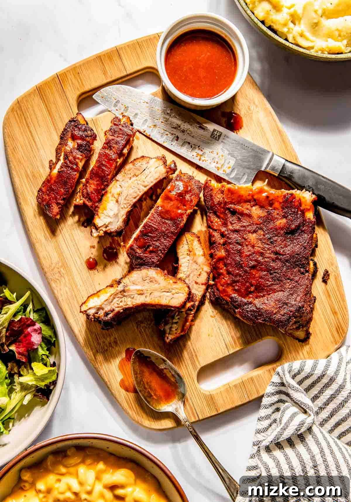 Fall-Off-The-Bone Oven Ribs 2 Sliced baby back ribs on a wooden board with a knife, small bowl of sauce, and spoon, surrounded by dishes of mashed potatoes and salad.