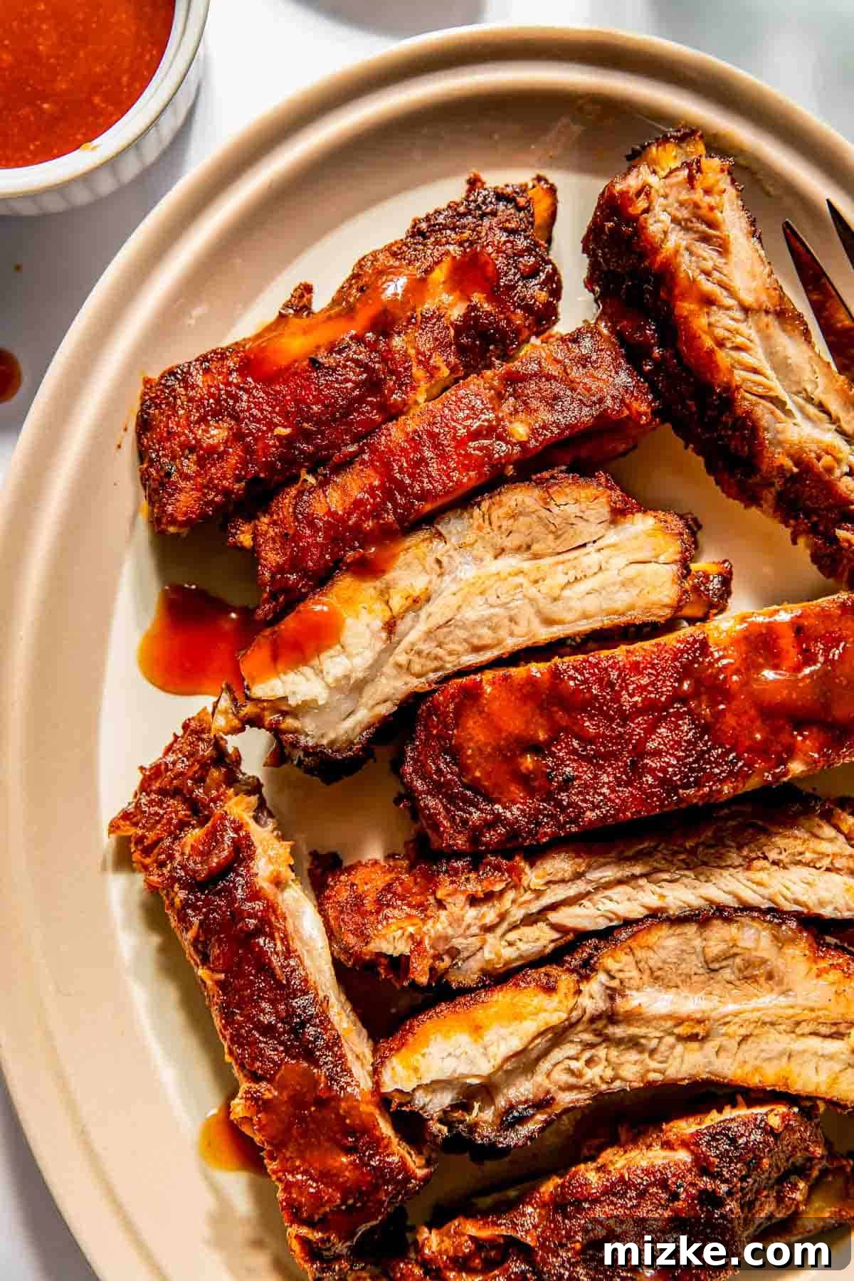 Fall-Off-The-Bone Oven Ribs 3 Close-up of sliced, seasoned baby back ribs on an oval plate with barbecue sauce. A small cup of sauce is visible in the background.