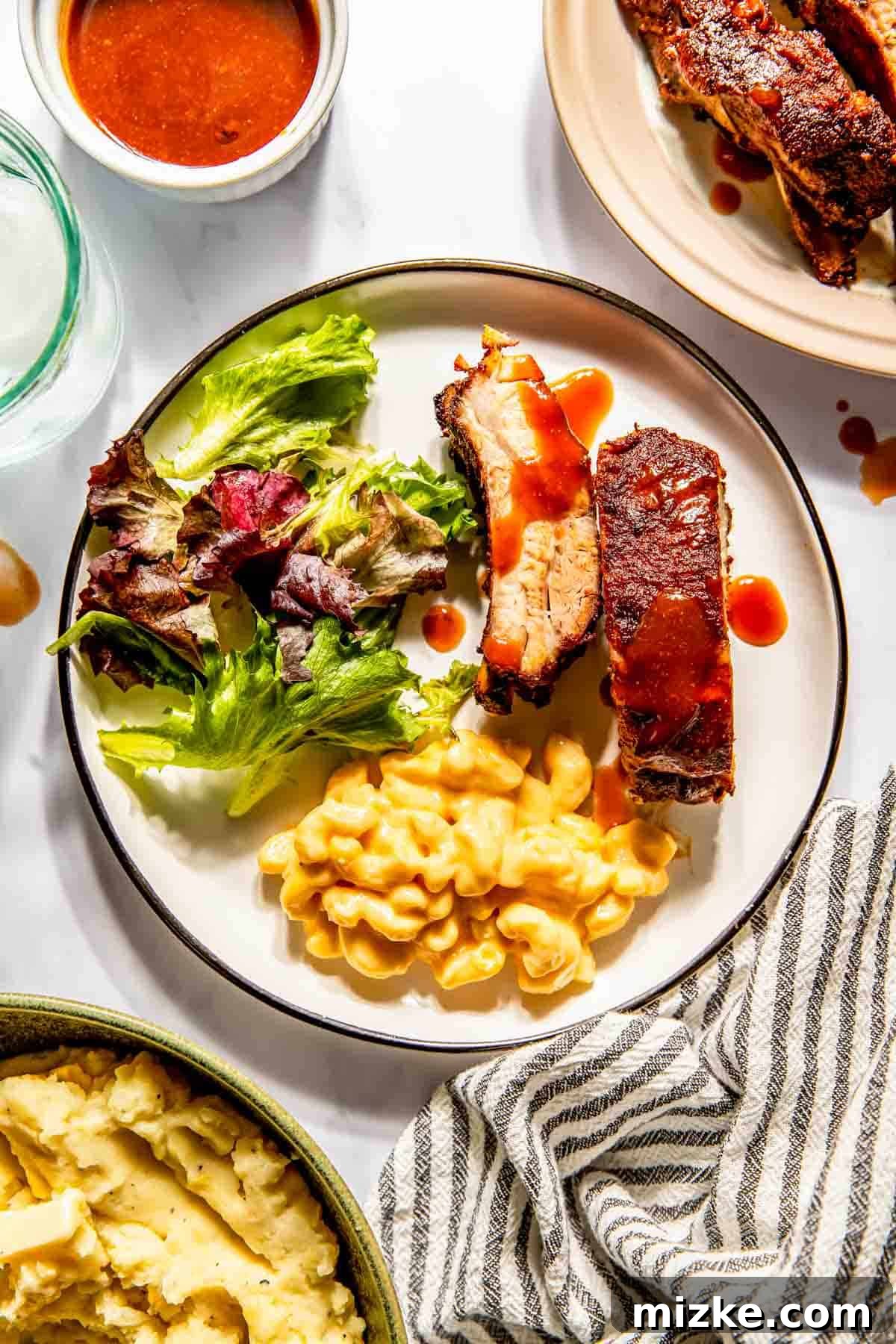 Fall-Off-The-Bone Oven Ribs 4 Plate of baby back ribs with sauce, accompanied by macaroni and cheese and a side salad. Nearby are mashed potatoes and a small bowl of extra sauce.