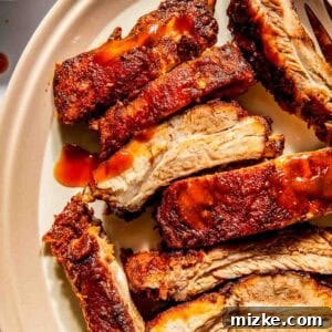 Close-up of sliced, seasoned baby back ribs on an oval plate with barbecue sauce. A small cup of sauce is visible in the background.