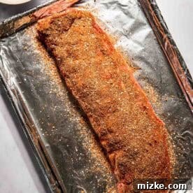 Raw rack of ribs seasoned with spices on a foil-lined baking sheet, next to a bowl of seasoning.