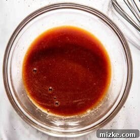 A glass bowl containing a bbq sauce ingredients with a whisk beside it on a white surface.
