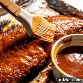 Brushing barbecue sauce on a rack of ribs on foil, with a small bowl of sauce nearby.
