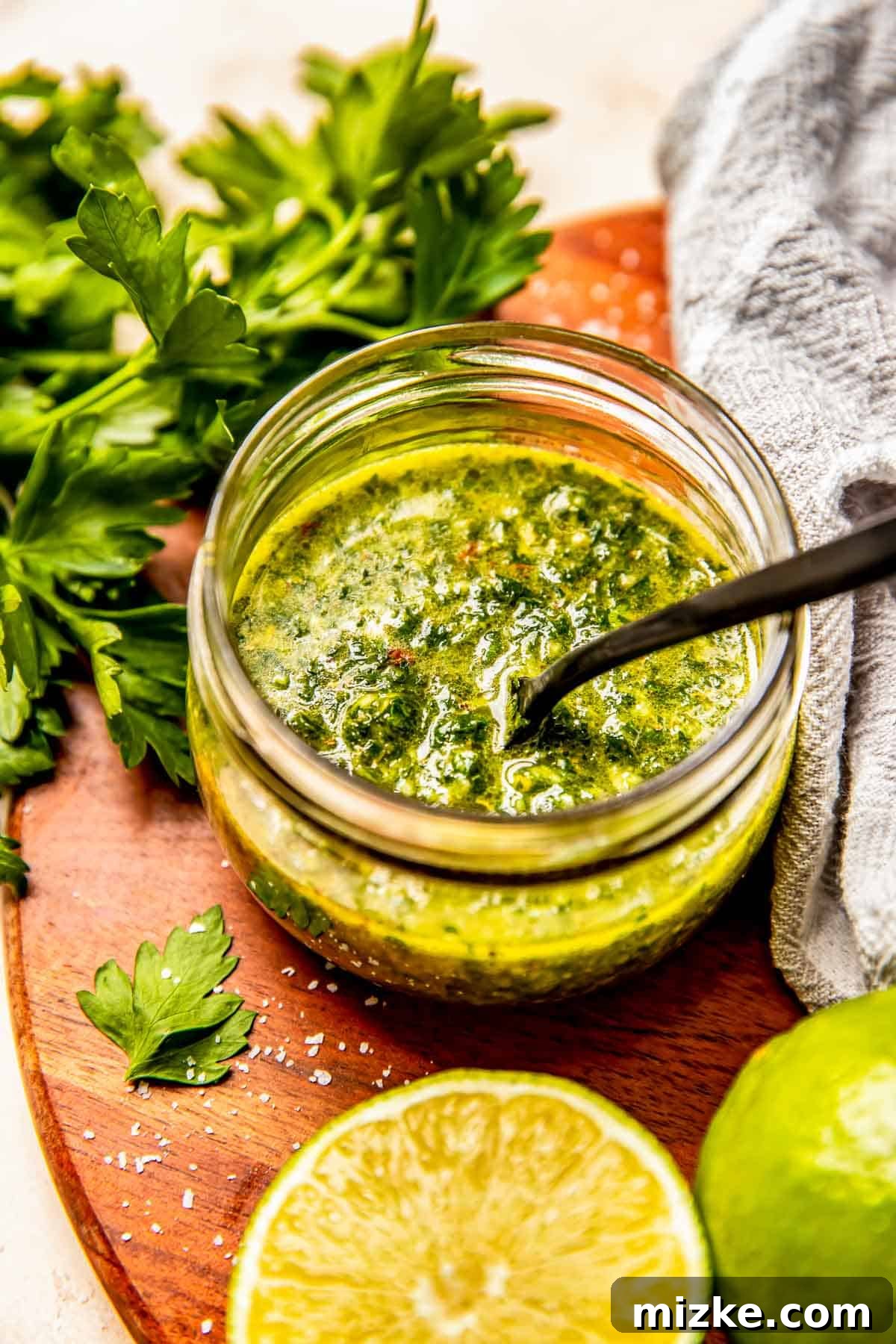 chimichurri sauce in a small glass jar with a black spoon. the jar is sitting next to fresh parsley, limes, and a linen towel on a wooden cutting board