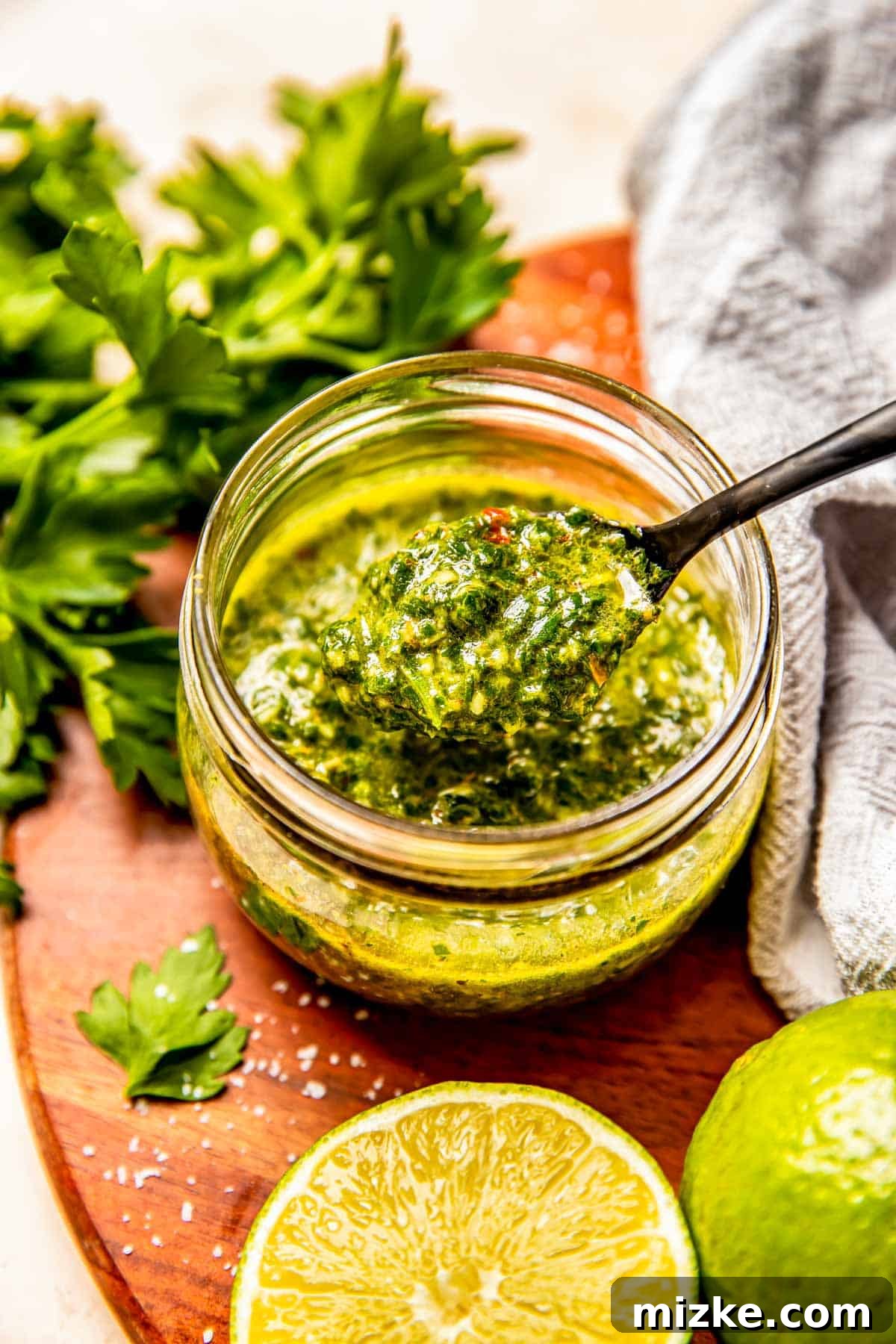 a black spoon has a scoop of chimichurri sauce scooped up from inside a glass jar
