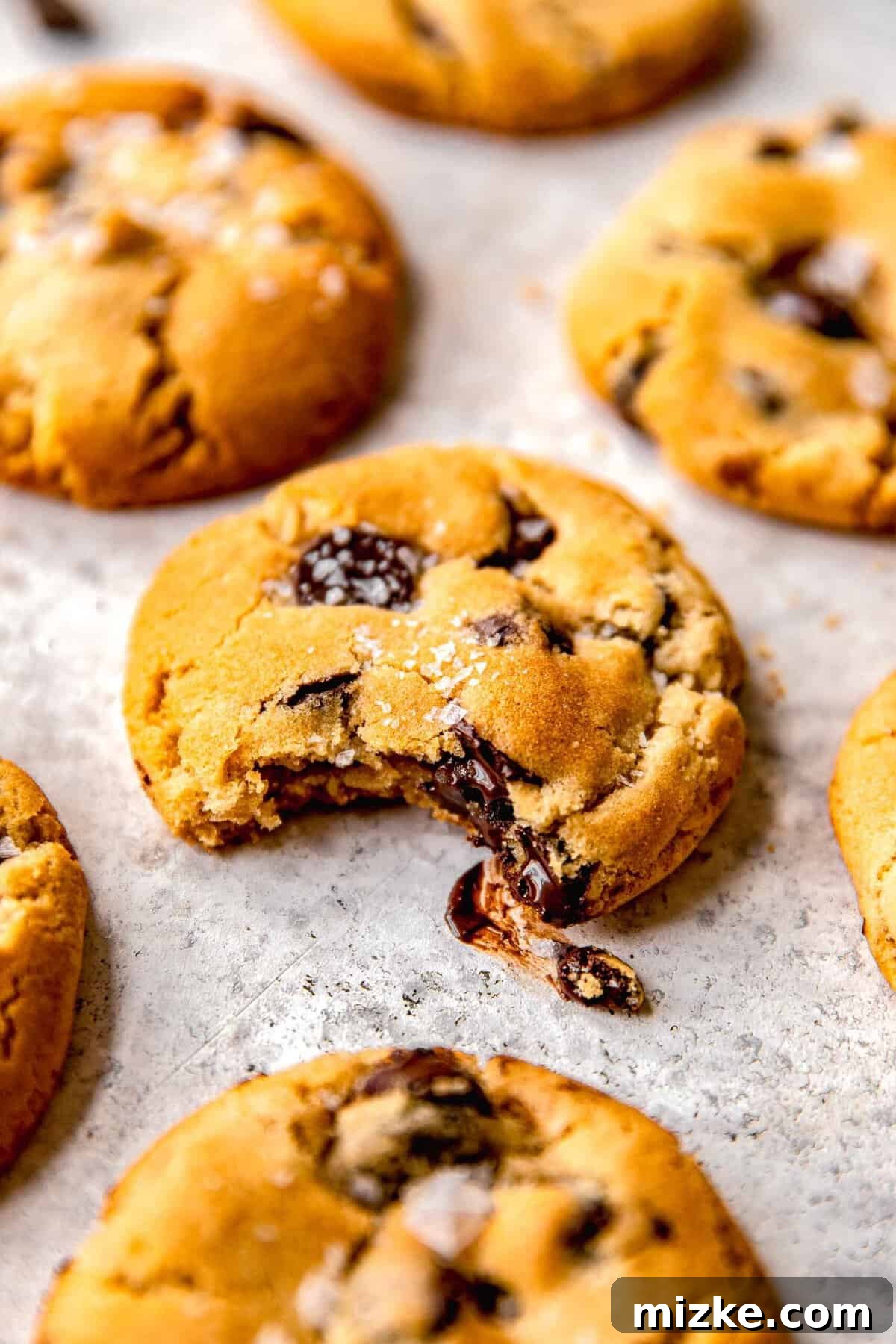 Close-up of a warm chocolate chip cookie with a bite taken out, sitting on a baking sheet, showing its gooey center.