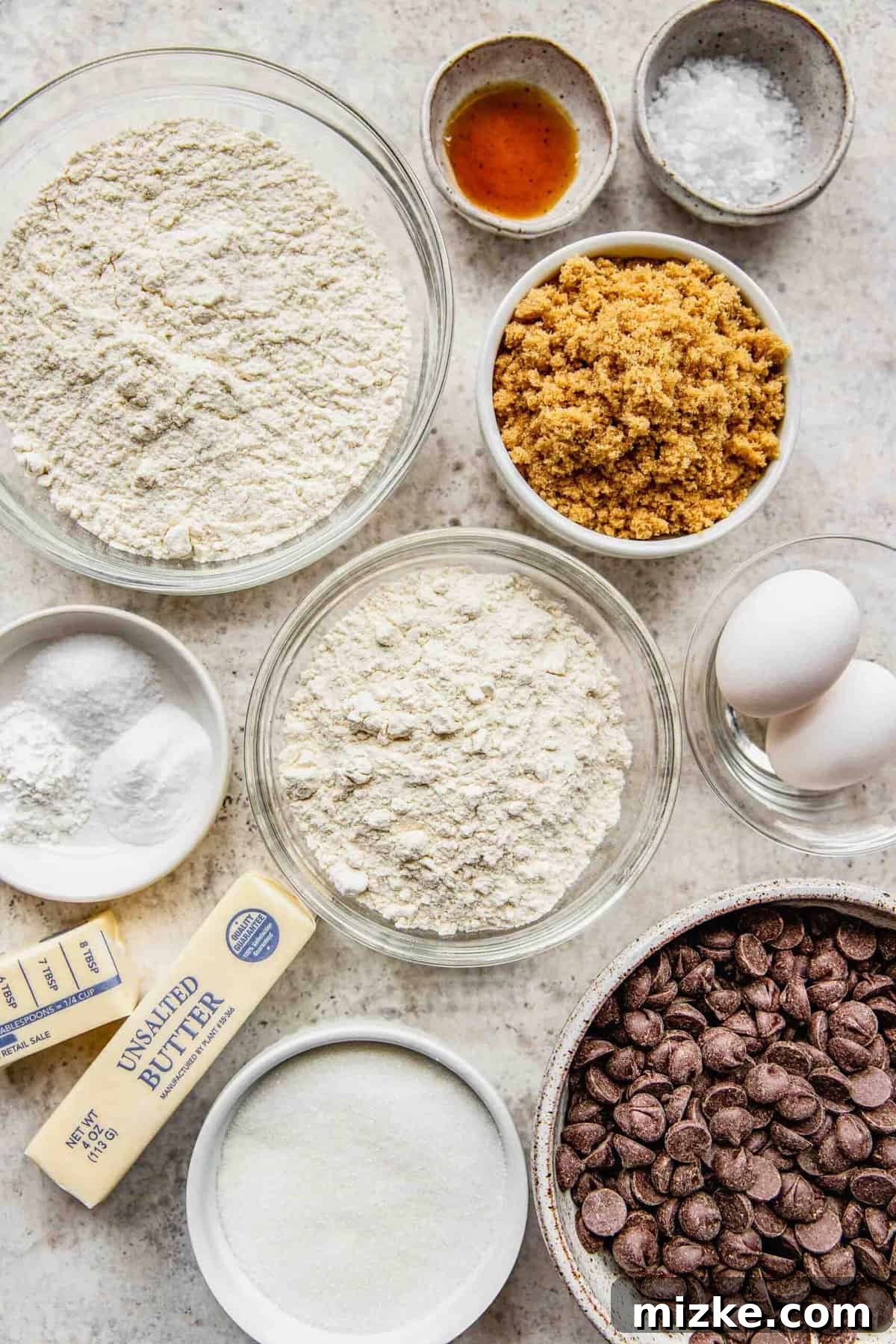 A curated display of ingredients for chocolate chip cookies, including flours, sugars, butter, eggs, vanilla, and dark chocolate chips.