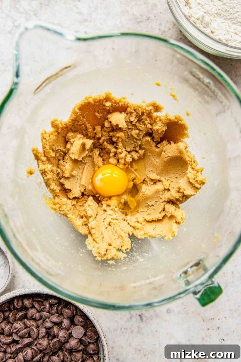 Adding a single large egg into the creamed butter and sugar mixture, preparing to mix for the cookie dough.