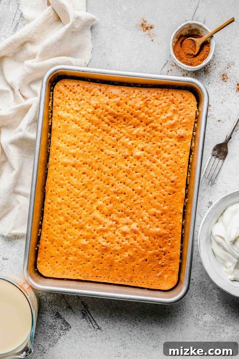 Cake with holes poked, ready for the three-milk soak. Cooled yellow cake in a pan with fork holes, next to a pitcher of milk mixture, Cool Whip, and cinnamon.
