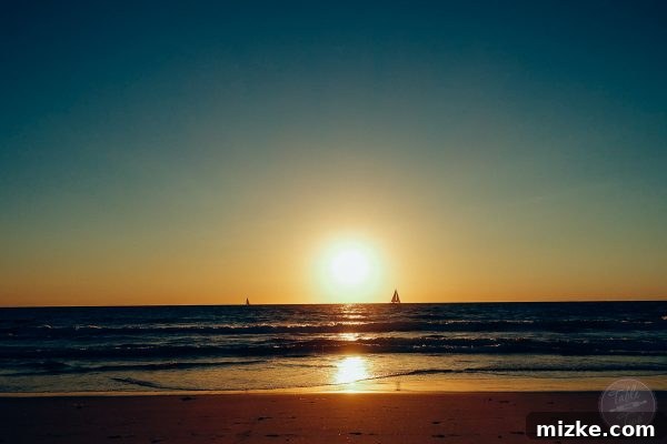 Spectacular sunset over the Pacific Ocean, with sailboats on the horizon and palm trees silhouetted.