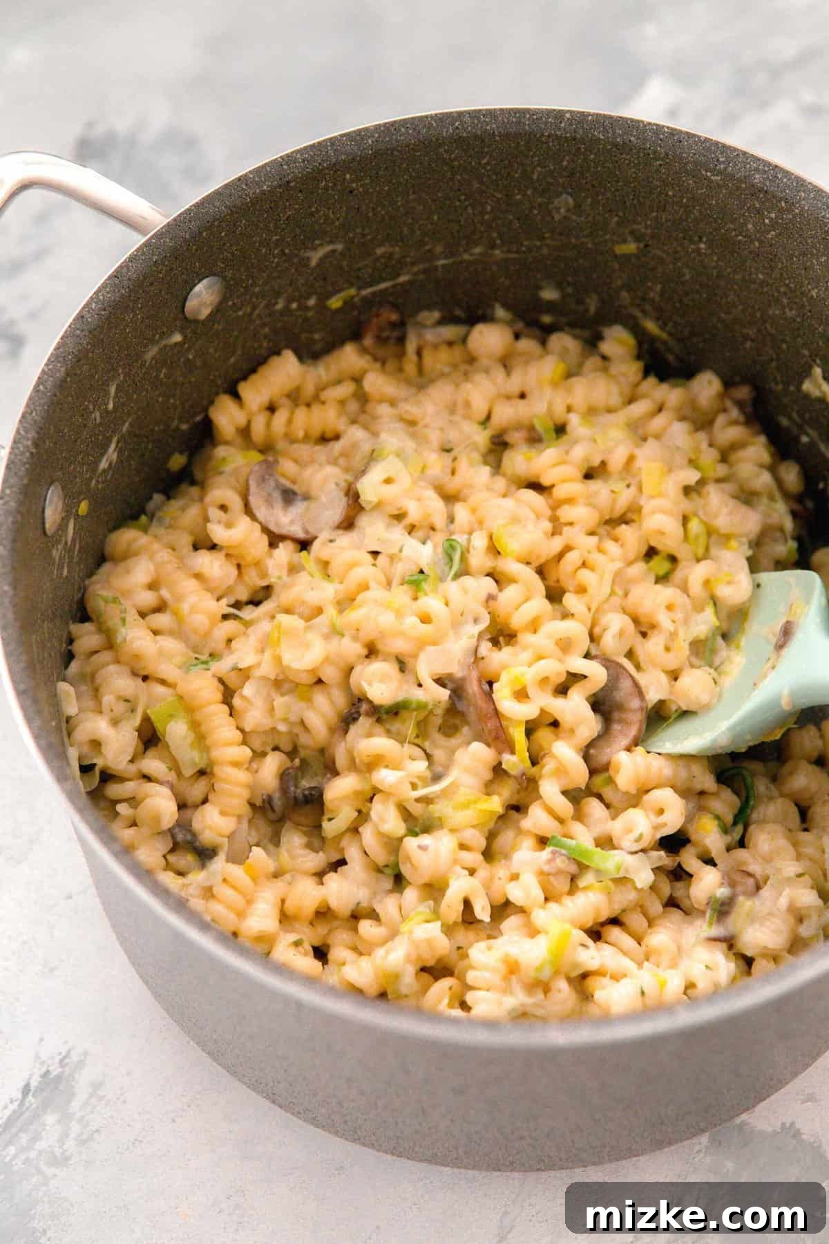 Velvety Mushroom and Leek One-Pot Pasta 11 Creamy leek and mushroom pasta being gently tossed with melted Boursin cheese in a large pot using a mint green silicone spatula.